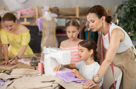 Teenage girl tries to sew on sewing machine. Teacher helps. Children watch and learnの写真素材