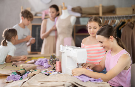 Teenage girl works on sewing machine while teacher and children learn how to cut fabric