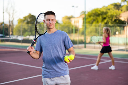 Portrait of positive young man with racket and ball in his hands on tennis court outdoorの写真素材