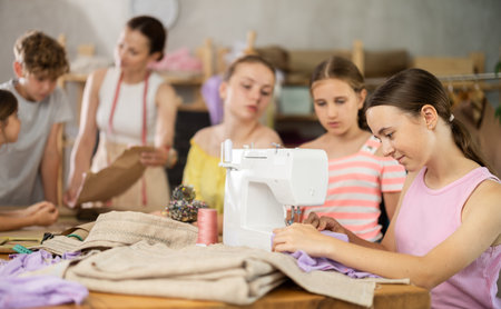 Teenage girl works on sewing machine while teacher and children learn how to cut fabricの写真素材