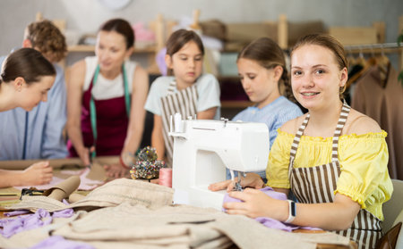 Teenage girl works on sewing machine while teacher and children learn how to cut fabric