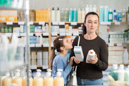 Woman with schoolgirl daughter are busy choosing cream at pharmacyの写真素材