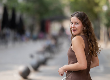 Back view of cute young girl wearing stylish short summer dress with white handbag walking along shady tree lined street enjoying spring day in Barcelonaの写真素材