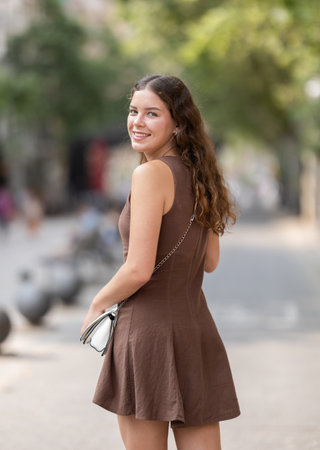 Back view of beautiful stylish young girl in dress with white handbag posing on city street on summer dayの写真素材