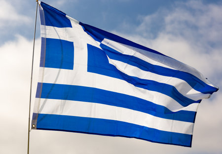 Against background of cloudy sky,white-blue flag of Greece flutters on flagpoleの写真素材