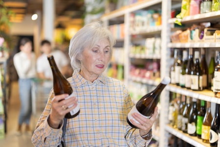 Senior female customer in supermarket examines bottles of wine, chooses alcoholic drink.の写真素材