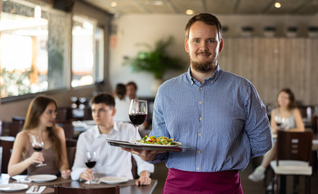 Professional friendly waiter holding serving tray with glass of wine for restaurant guestsの写真素材