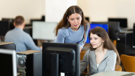 During computer lesson, female teacher helps girl student complete difficult taskの写真素材