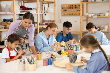 Children paint ceramic work-pieces at table with teacher in art studioの写真素材