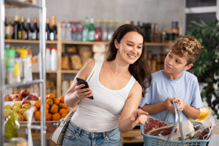Mom chooses products from a list on her smartphone. Teenage son helps carry shopping cart in supermarketの写真素材