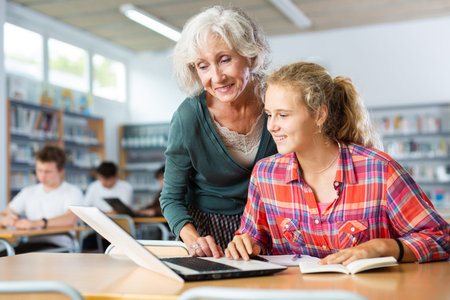 Mature female teacher working with schoolgirl in computer class of library