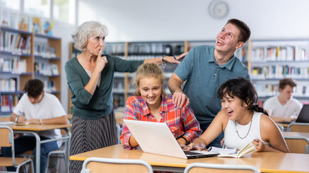 Teacher shows silence sign to laughing schoolchildren teenagers while working on laptop
