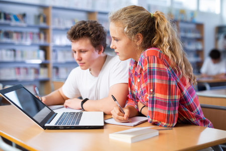 Positive female and male students teenagers study together at laptop while sitting in school library