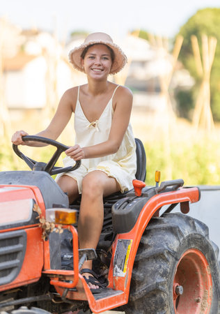 Young woman sits on minitractor and drives rural vehicle, performs loading work on farm