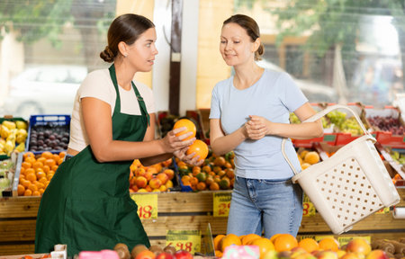 Polite salesgirl offering sweet oranges to female customer in greengrocery store