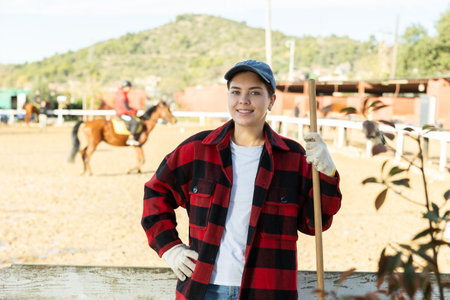 Smiling skilled young woman in plaid shirt smiling at camera while holding working tool on horse ranch in springの写真素材