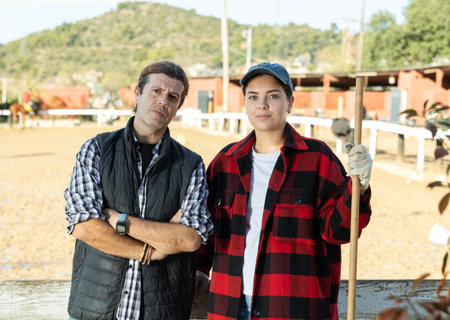 Experienced man and woman in plaid shirts looking at camera while holding working tool on horse ranch in springの写真素材