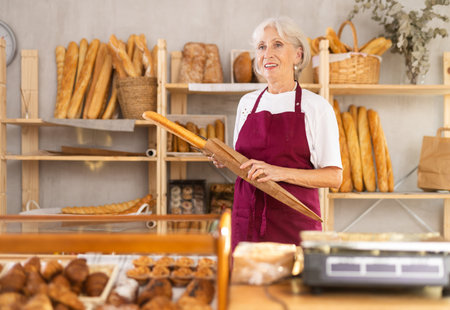 Mature saleswoman holding paper bag with baguettes in bakery
