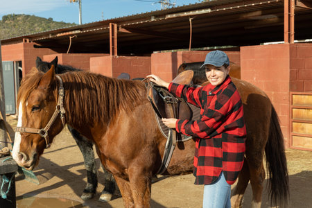 Portrait of positive young female worker in plaid shirt standing close to saddled horse in...
