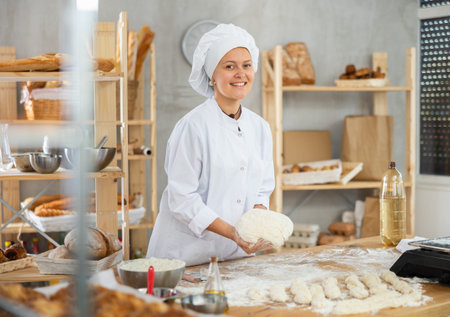 Young baker girl is standing near work table in kitchen, working with dough