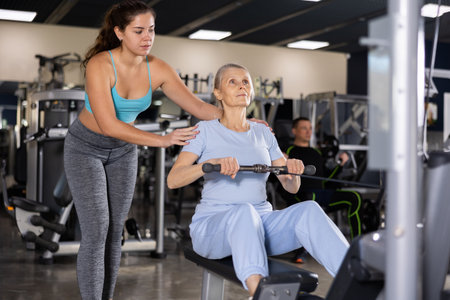 Girl coach helping elderly woman exercising back on cable machineの写真素材