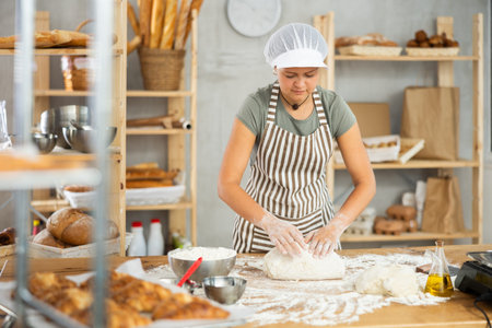 Young smiling female baker preparing raw yeast dough on kitchen table in bakery