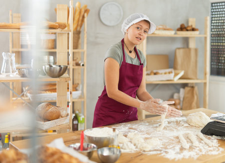 Female baker prepares raw dough in bakery, forming it into long sausages to be cut into pieces for baking buns or croissantsの写真素材