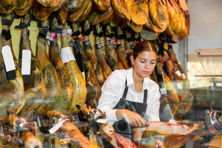 Female seller cutting slices from whole leg of jamon in meat store