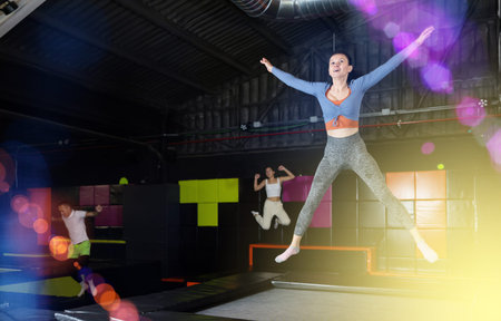 Positive middle aged woman in sporty clothes jumping vertical high in modern trampoline hall