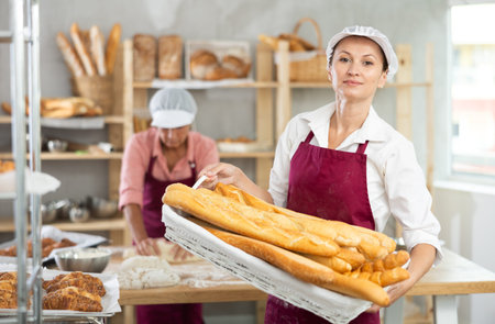 European young bakery female stands proudly in front of fresh bread holding basket of freshly baked...