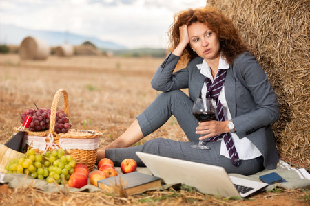 Tired business woman in stylish gray suit with white shirt and loosened tie sitting on blanket with picnic basket and laptop, leaning against bale of straw on mown field, relaxing with wine in retreatのeditorial素材