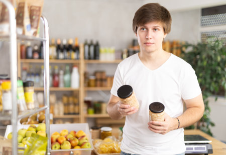 Young man store visitor chooses can of beans, hold and view glass jar package.