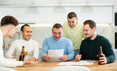Happy friends drinking beer while sitting together at home cooking table