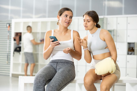 In locker room of sports club, girl communicates with female friend holding phone in her hands.
