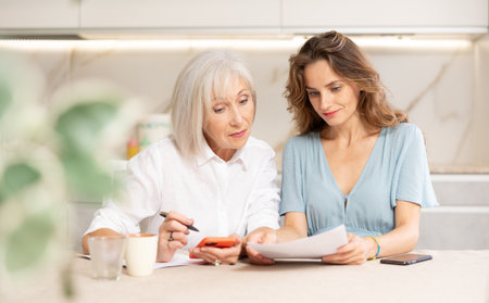 Mother and daughter sitting at table and calculating finances. Daughter using smartphone for online paymentの写真素材
