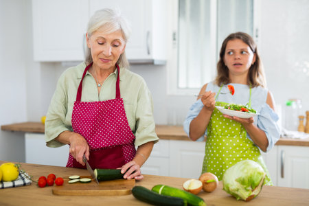 Elderly mother is preparing dinner in the kitchen. Adult daughter tries salad in backgroundの写真素材