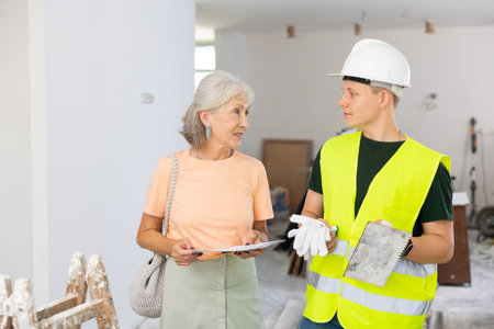 Mature woman discussing room renovation work plan with guy in protective helmet and yellow vestの写真素材