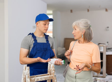 Young builder in blue overalls and an elderly woman are discussing the renovation of room in an apartmentの写真素材