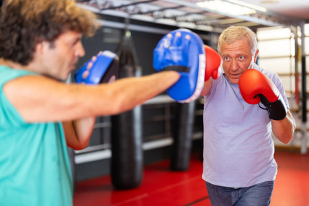 Mature hobby boxer practicing punches on mitts with instructor