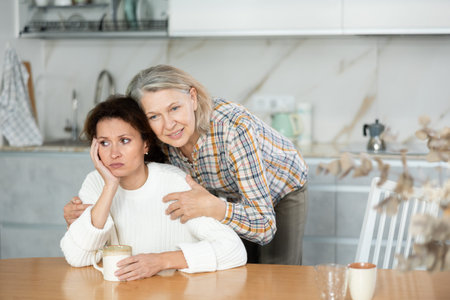 Middle-aged woman sitting at the kitchen-table and old woman trying to calm her down