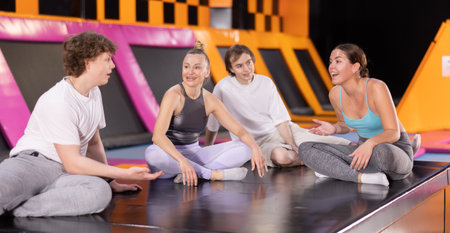 Group of enthusiastic young people sitting on trampoline and discussing upcoming jumping workout in entertainment centerの写真素材