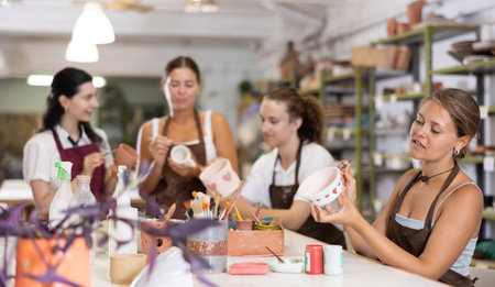 Young woman sits near table and decorates clay ceramic craft vase, plateの写真素材