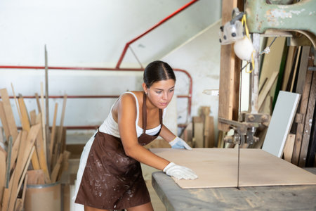 Girl employee of workshop cuts wooden board into parts using sawの写真素材