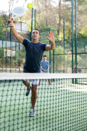 Young man serving ball while playing paddle tennis on outdoor court