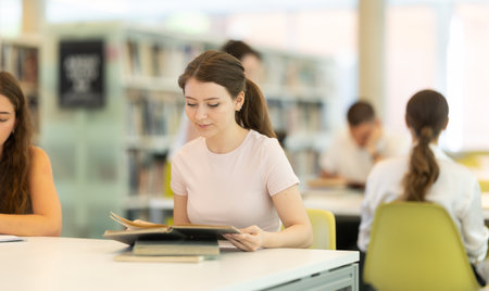 Female student sits at table in library and studies, reads books and takes notesの写真素材