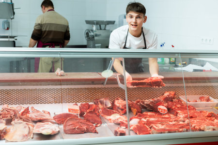 Young guy stands behind counter in meat department of supermarket and and holds piece of raw meat in...