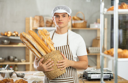 Happy male baker posing with basket of freshly baked baguettes in bakery, demonstrating concept of food industry and baker professionの写真素材