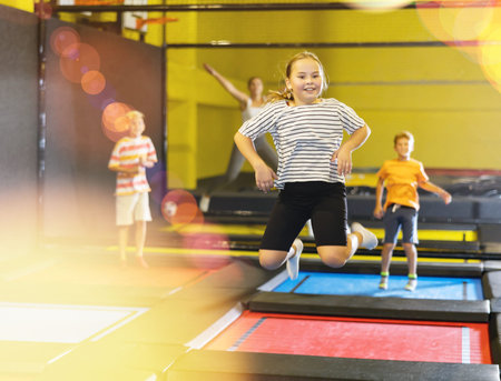 Smiling school-age girl in striped T-shirt and black shorts jumping and indulging on trampolines in entertainment center. Active leisure conceptの写真素材