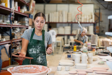 Young female worker in pottery factory lowers fired clay plates and cups into container with glaze