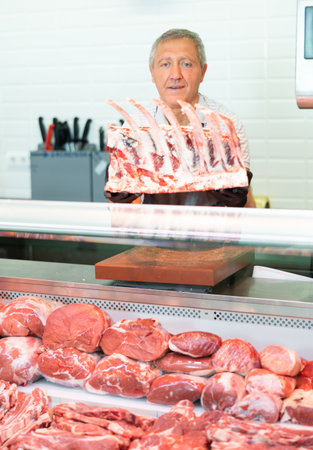 Adult man seller lays out beef ribs in butcher shopの写真素材